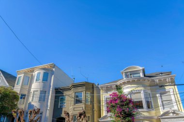 Tangled electrical wires against the row of houses in San Francisco, CA. Three buildings with gray house in the middle of flat apartment on the left and yellow house on the right with curved wall.