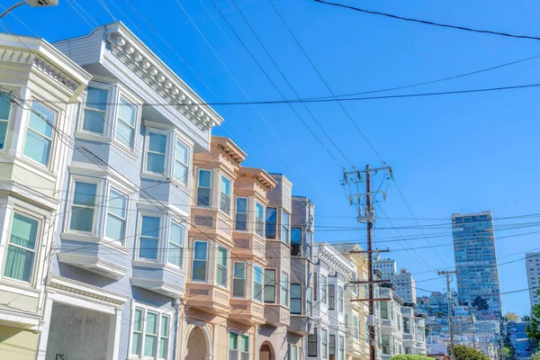 Row of flat apartments with pastel colors in San Francisco, CA. There are electrical posts and wires at the front of the apartments and a view of high-rise building and sloped street at the back.