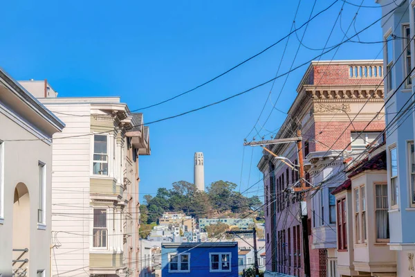 View of a tower on top of a hill from a neighborhood with victorian houses at San Francisco, CA. There are houses on both sides with electrical wires in the middle against the building at the back.
