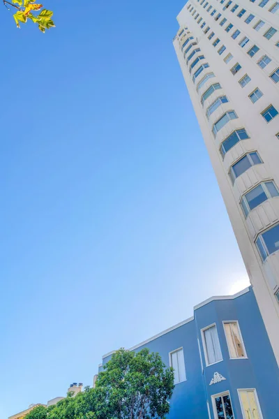 Low-rise flat apartment beside the high-rise building in San Francisco, CA. There is a blue house near the tree on the left beside a tall apartment with bay windows against the sky at the background.