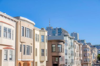 Row of apartments in San Francisco, CA near the commercial buildings at the back. There are flat apartments at the front and a view of taller buildings at the back against the clear sky background.