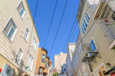 Electrical wires in the middle of rows of houses in a low angle view at San Francisco, CA. Residential buildings with wood sidings and a view of tall building at the back.