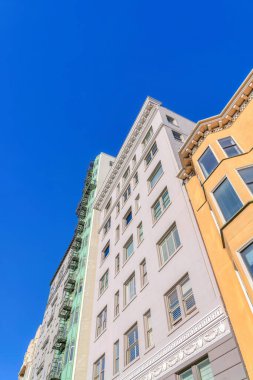 Multi-storey residential buildings in a low angle view at San Francisco, CA. There is a mid-rise gray building at the center of yellow and green buildings against the clear blue sky.