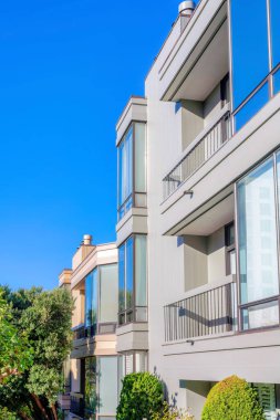 Row of three-storey houses with balconies beside the reflective picture windows in San Francisco, CA. There are trees and bushes at the front of the houses with two balconies on the right.