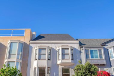 Facade of adjacent houses with bay windows against the blue sky at San Francisco, CA. There is a house on the left with roof deck along with the houses with roof shingles and gray wall exterior.