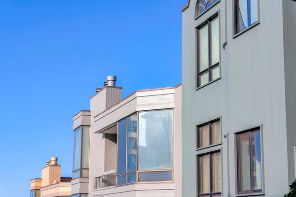 Row of houses with balconies, picture windows, and flues against the sky in San Francisco, CA. There is a gray building on the right with paned windows near the houses with reflective windows.