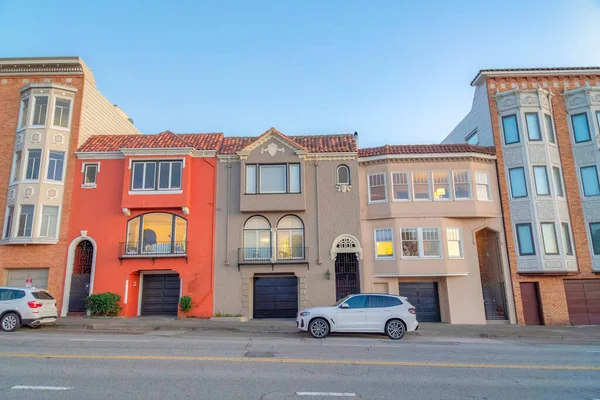 Rowhouses in San Francisco, California with metal gate doors and attached garages. There are apartments on both sides with bricks and bay windows near the street with parked vehicles on the side.