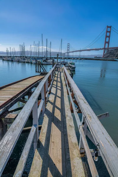 Wooden pier with handrails heading to the boats against the Golden Gate Bridge in San Francisco, CA. Narrow path of a pier with light bulbs on its handrails with a view of the bridge and mountains.