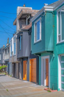 Small suburban houses with attached garages and view of electrical wires above in San Francisco, CA. There is a concrete pavement at the front of colorful houses with different wall sidings.