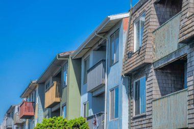 Side view of houses with two balconies in San Francisco, CA. There is a house at the front with wood shingle sidings along with the houses with painted stucco walls against the clear blue sky.