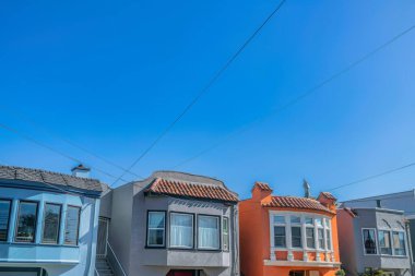 Facade of adjacent small houses with stairs at the entrance on the side at San Francisco, California. Colorful exterior of houses with electrical wires above against the clear blue sky.