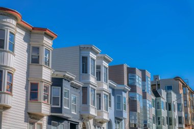 Side-view of rowhouses with bay windows against the sky in San Francisco, California. Facade of houses with different kinds of exterior and painted wood sidings and stucco walls.