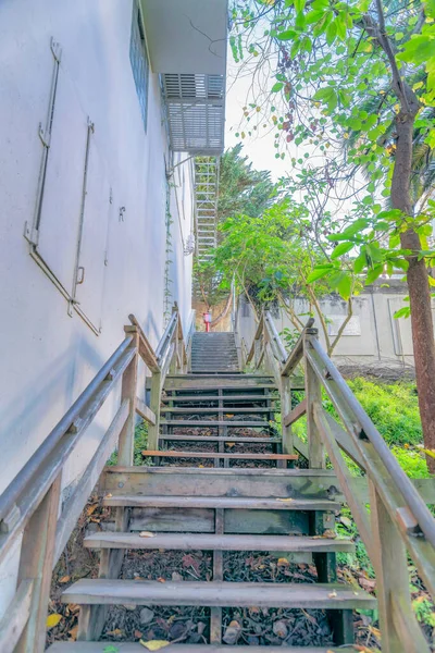 Wooden outdoor narrow staircase beside a residential building in San Francisco, CA. There is a building on the left beside the staircase on a slope with trees and wild plants on the side.