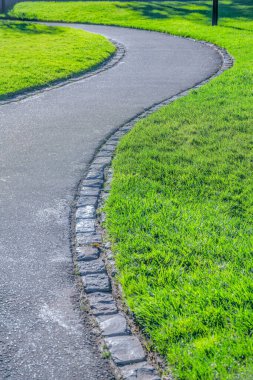 Curved asphalt trail with stone trims on the side in San Francisco, California. Concrete walkway outside in the middle of the green grass lawn.