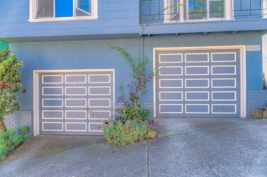 Attached garage of townhomes with identical garage doors on a sloped neighborhood- San Francisco, CA. Two sectional garage doors below the window on the left and balcony on the right.