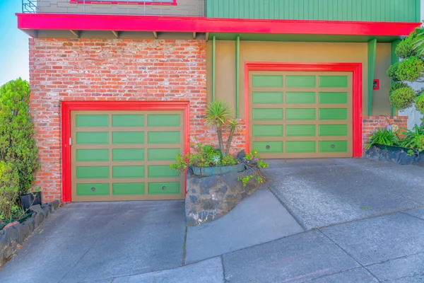 Two garage doors on a slanted concrete street at San Francisco, California. Two townhouses with the same garage doors, red trims, and brick walls on the left and painted stucco wall on the right.