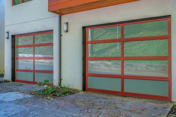 Modern garage exterior with two garage doors and wall lights in San Francisco, California. There are two garage doors with reflective window panels and a concrete driveway with scattered dried leaves.