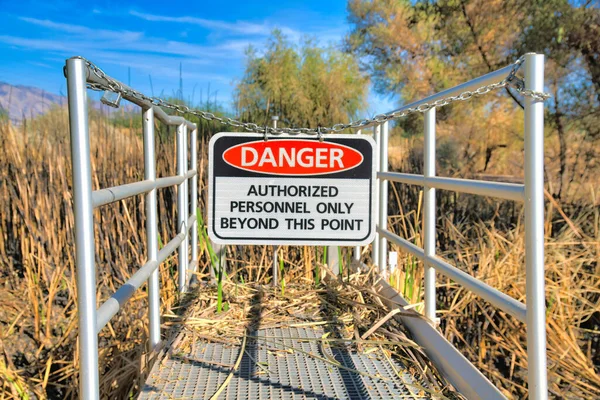 Hanging danger sign on a chainlink at Sweetwater Wetlands in Tucson, Arizona. Danger Authorized Personnel Only Beyond This Point sign on a metal deck against the dried grass at the back.