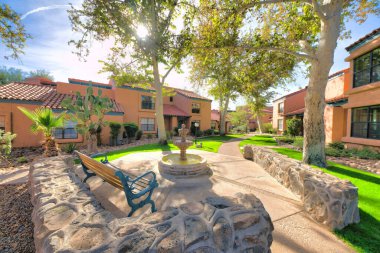 Bench near the small fountain outside the residences with italianate design exterior in Tucson, AZ. Lounge area with low stone walls near the houses with lawn and lamp post at the entrance.