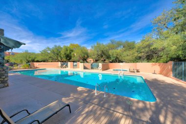 Clubhouse pool in a residential area at Tucson, Arizona. There is a lounge chair on the left on a concrete pavement near the pool with jacuzzi pool near the painted wall with railings at the back.