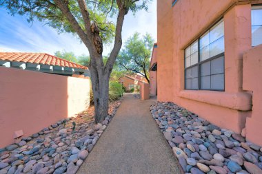 Rough concrete walkway with pebbles and tree on the side near the houses in Tucson, AZ. There is a wall with a view of clay tile roof across the houses on the left with the same peach painted walls. clipart