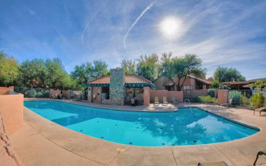 Clubhouse pool with lounge chairs and dining area with stone walls at Tucson, Arizona. Public pool with painted walls and railings against the trees and houses at the back.