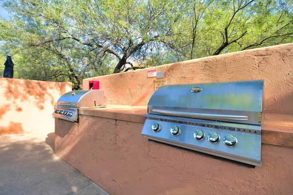 Two gas barbecue grills on a concrete deck outside in Tucson, Arizona. Deck mounted grills with a signages on a peach concrete wall at the middle against the trees at the background.