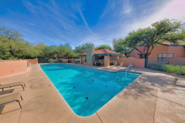Clubhouse pool near the houses in a residential area at Tucson, Arizona. There are lounge chairs near the pool and a view of a building with stone walls and clay tile near the house on the right.