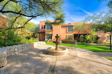 Small water fountain and wooden bench against the peach italiante houses at the back in Tucson, AZ. There is a low stone wall on the left near the trees and a lawn near the entrance of the houses.