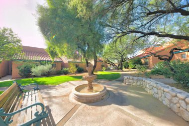 Small lounge area with bench and fountain outside the houses with peach exterior in Tucson, AZ. There is a wooden bench on the left and stone wall on the right against the view of houses and lawn.
