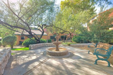 Water fountain and bench in a neighborhood at Tucson, Arizona. There are large trees on the side of walkways with lamp post near the houses at the back.