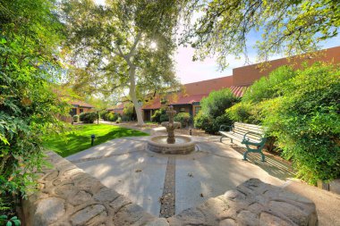 Water fountain and bench surrounded by the italianate houses in a subdivision at Tucson, Arizona. There is a stone wall and plants near the wooden bench on the right against the houses at the back.