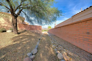 Dirt path with stones on both side near the trees and wall bricks at Tucson, Arizona. Walkway with a view of trees on the left and walls near the houses on the left and right sides.