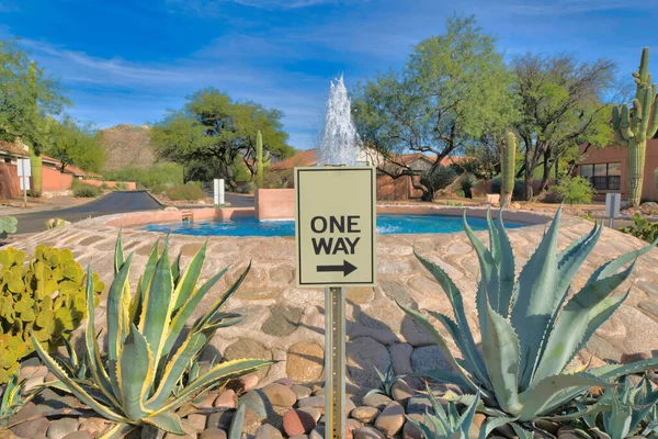 One way signage in between large succulent plants near the large water fountain at Tucson, Arizona. One way rotational road in a residential area near the mountains.