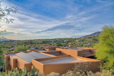 Top of a medterranean house with solar panel and clay tile roofs at Tucson, AZ. There are bunny ear cactuses and wild shrubs at the front and a view of sloped neighborhood near the mountains.
