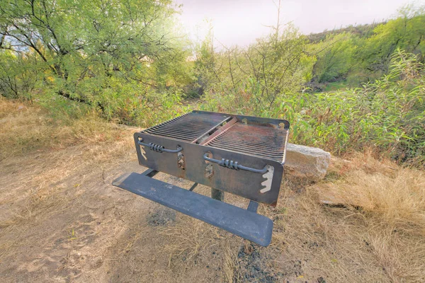 Post-mounted campground charcoal fire box with two grills in Tucson, Arizona. Barbecue grills on a campground with a view of a shrubland and sunset at the back.