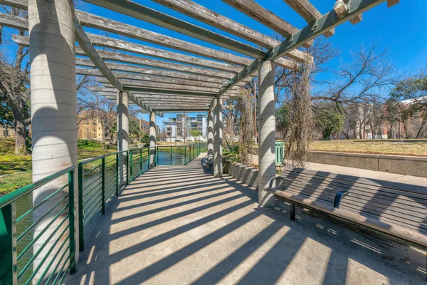 Concrete walkway with pergola and bench near the river at San Antonio, Texas. There is a railings on the left from the river and bench on the right against the field and buildings at the background.