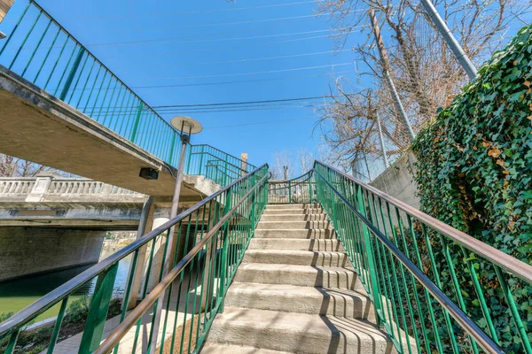 Staircase with metal railings near the river at San Antonio, Texas. Concrete steps leading to a bridge with lamp post on the left near the ramp and a view of a wall with crawling plants on the left.