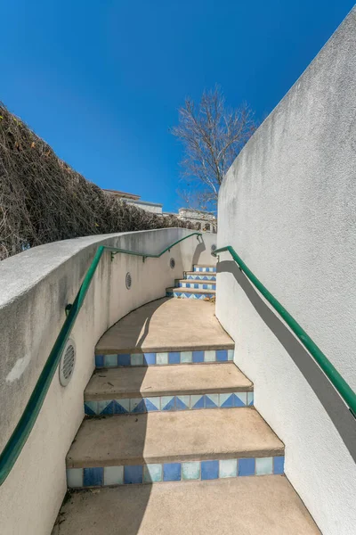 Outdoor stairs with blue tile risers and wall-mounted green handrails at San Antonio, Texas. Stairs in between concrete walls and a view of vines on the left and building at the back against the sky.