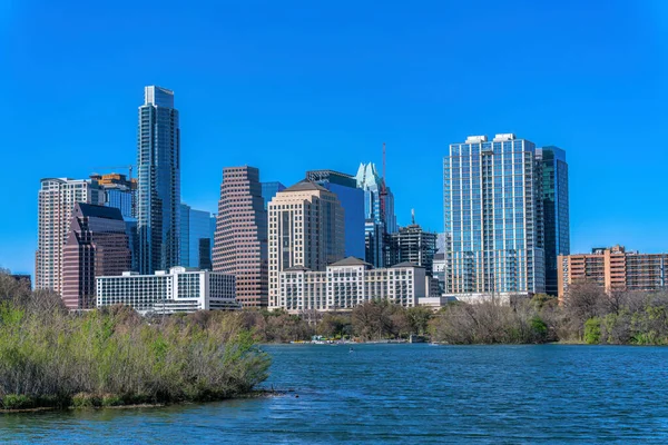 Austin, Texas cityscape and view of Colorado River at the front. There are trees at the shore of the river near the buildings against the clear blue sky at the back.