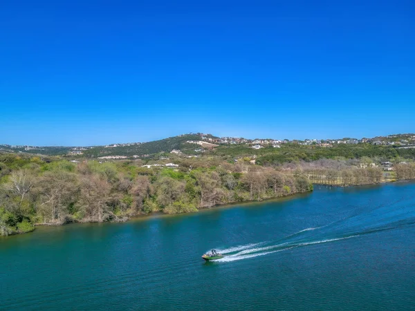Austin, Texas- Speed boat passing through the Colorado River. High angle view of a forest park and mountain with buildings on top against the clear blue sky.