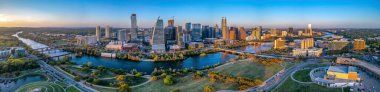 Panoramic cityscape of Austin, Texas near the Colorado River against the sunset skyline. There are parks at the front near the river and a view of skyscraper buildings across.