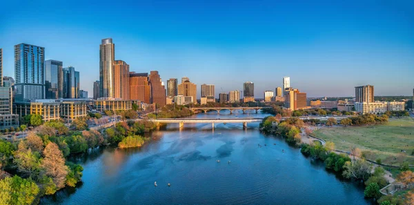 Colorado River near the district urban area of Austin, Texas. There are bridges over the river and a view of skyscraper buildings on the left and large field at the right side against the sunset sky.
