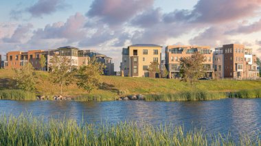 Panorama Puffy clouds at sunset Oquirrh Lake at the front of residential complex buildings at Daybreak, Utah. Reflective lake waterfront with grasses on the shore and arch bridge on the left
