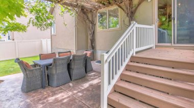 Panorama Outdoor patio with wooden pergola roof with vines and string lights. There is a sliding glass door with wooden doorsteps and railings near the dining table with woven chairs