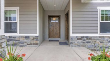Panorama Porch of a house with potted flowers and two wooden front doors. Exterior of a house with gray vinyl wood and stone veneer sidings.