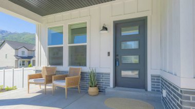 Panorama Porch with two woven armchairs and snake plant on a woven pot. Entrance exterior with black front door with glass panels and lockbox near the windows and a view of the neighborhood