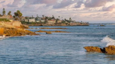 Panorama Puffy clouds at sunset Coastal residential area at La Jolla in California. Buildings near the pacific ocean on the right with rocks and concrete seawalls on the left.