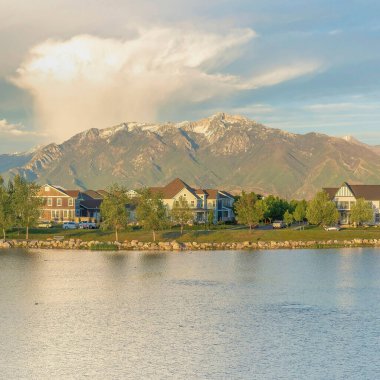Square view of the residential area against the mountain range background at Daybreak, Utah. Oquirrh lake waterfront with field and rocks near the shoreline and water with reflection of the sky.