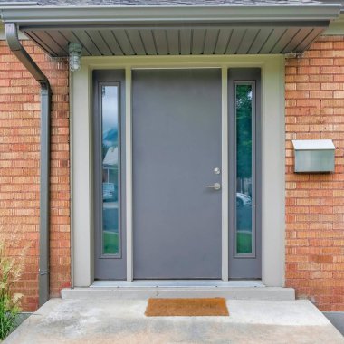 Square Modern gray front door with two side panels and mailbox on the side. Home exterior with bricks and stairs at the doorsteps in the middle of the tall grass on the side.